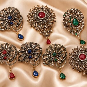 May include: A collection of seven ornate metal brooches featuring antique gold-tone finishes, intricate filigree patterns, and embedded clear crystals. Each brooch is accented with a central colored gemstone and a matching teardrop-shaped pendant.