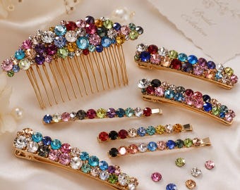 May include: A collection of gold-tone hair accessories including one large hair comb and five hair clips, all embellished with multi-colored round rhinestones in shades of pink, blue, green, yellow, and clear, alongside several loose rhinestones.