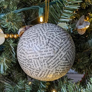 May include: A round Christmas ornament covered in text from a book, hanging on a green Christmas tree. The ornament is white with black text and has a gold string for hanging. The tree has small lights.