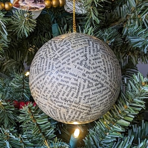 May include: A round Christmas ornament covered in printed text, hanging from a gold string. The ornament is white with black text and is hanging on a green Christmas tree with lights.