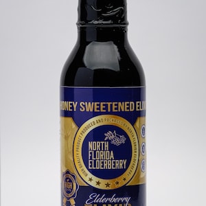 May include: A dark glass bottle of elderberry elixir with a black cap. The label features the text "Honey Sweetened Elixir" and "North Florida Elderberry" in gold and blue. The bottle is filled with a dark liquid.
