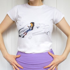 Puede incluir: Camiseta blanca con una ilustración en blanco y negro de una mujer conduciendo un coche.