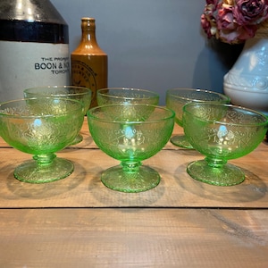 Può includere: Sei coppette da dessert vintage in vetro verde con un motivo floreale. Le coppette hanno una base a piedistallo e una forma arrotondata. Il vetro ha una leggera lucentezza e le coppette sono disposte su una superficie di legno.