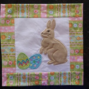 May include: A white fabric square with a brown bunny and two colorful Easter eggs embroidered on it. The fabric is surrounded by a green, pink, and white patterned border.