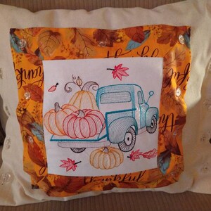 May include: A white pillow with a colorful embroidered design of a blue truck carrying pumpkins and fall leaves. The truck is surrounded by a border of orange and yellow leaves. The text "Happy & Thankful" is embroidered on the pillow.