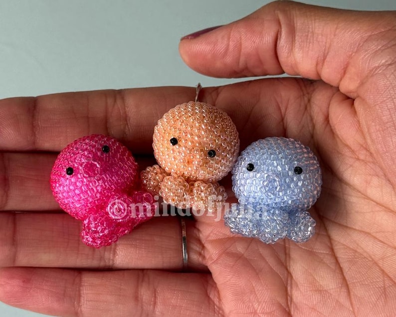 Pode incluir: Tr&ecirc;s pequenas figuras de polvo feitas de min&uacute;sculas contas transl&uacute;cidas em tons vibrantes de rosa choque, p&ecirc;ssego e azul claro. Cada polvo apresenta duas pequenas contas pretas redondas como olhos e uma cabe&ccedil;a esf&eacute;rica texturizada.