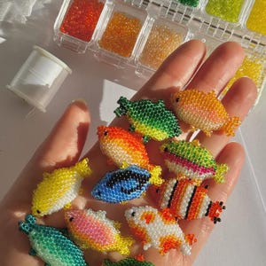 Puede incluir: Una mano sostiene una colecci&oacute;n de peces de cuentas de colores. Los peces son de varios colores, incluyendo amarillo, azul, naranja y verde. Peque&ntilde;os recipientes de cuentas de varios colores est&aacute;n en el fondo, junto con una bobina de hilo blanco. Los peces son hechos a mano.