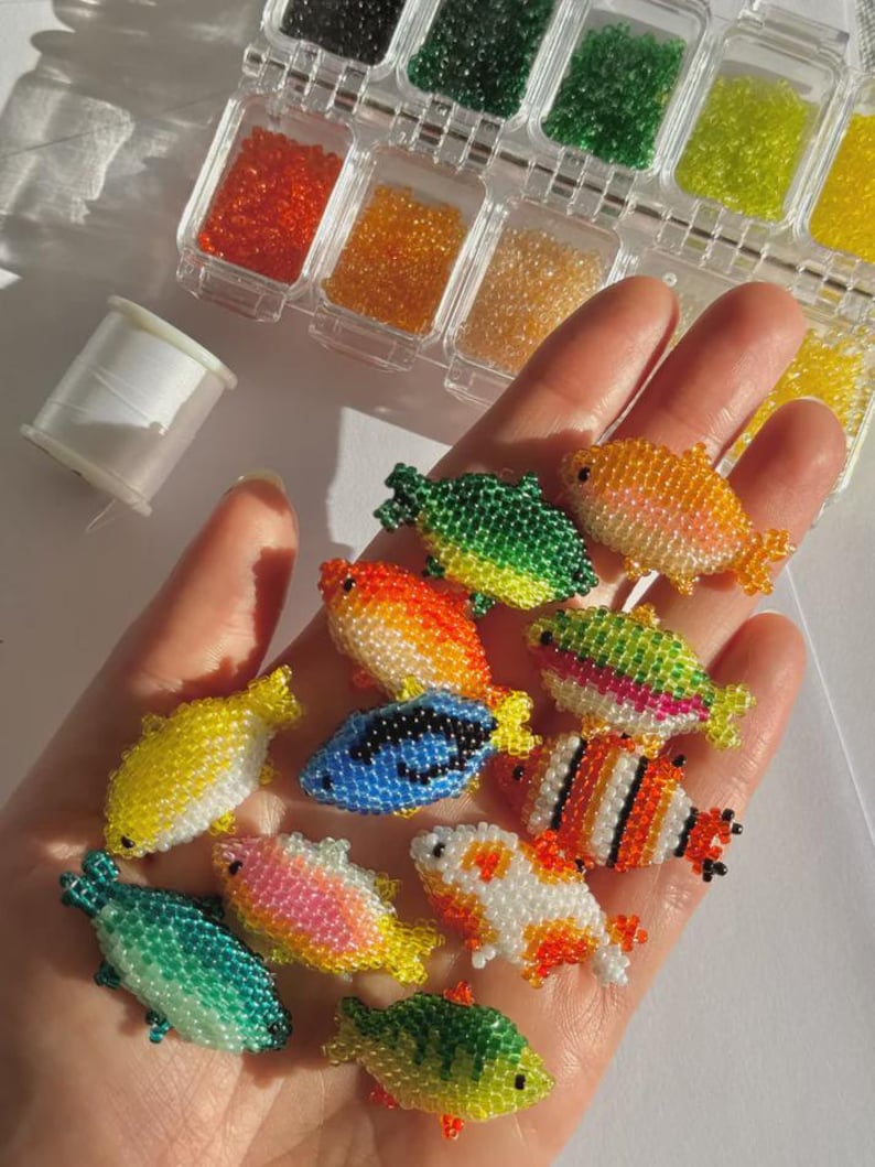 Puede incluir: Una mano sostiene una colecci&oacute;n de peces de cuentas de colores. Los peces son de varios colores, incluyendo amarillo, azul, naranja y verde. Peque&ntilde;os recipientes de cuentas de varios colores est&aacute;n en el fondo, junto con una bobina de hilo blanco. Los peces son hechos a mano.
