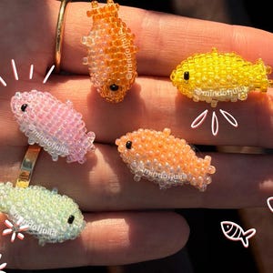 Pu&ograve; includere: Cinque pesci di perline nei toni del rosa, arancione, giallo e verde. Ogni pesce ha un occhio nero ed &egrave; fatto di piccole perline colorate. I pesci sono disposti su una mano, con un anello d'oro visibile.
