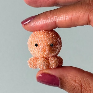 Pode incluir: Uma estatueta de polvo em miniatura feita de pequenas contas transl&uacute;cidas na cor p&ecirc;ssego. A criatura apresenta uma cabe&ccedil;a arredondada e v&aacute;rios tent&aacute;culos pequenos de contas na base, com duas min&uacute;sculas contas pretas s&oacute;lidas como olhos. O texto mindofjulia &eacute; vis&iacute;vel.