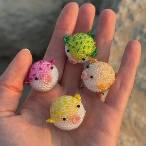 Puede incluir: Cuatro adornos de pez globo con cuentas en rosa, verde, amarillo y naranja. Cada uno tiene una barriga blanca y ojos negros. Los adornos se sostienen en una mano sobre un fondo borroso.