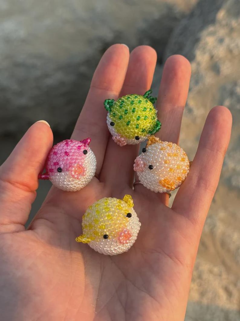 Puede incluir: Cuatro adornos de pez globo con cuentas en rosa, verde, amarillo y naranja. Cada uno tiene una barriga blanca y ojos negros. Los adornos se sostienen en una mano sobre un fondo borroso.