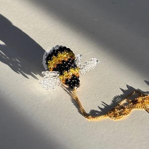 May include: A beaded bee pendant necklace. The bee is black, yellow, and clear, with a gold-colored chain. The necklace is on a white surface, with a long shadow cast by the sun.