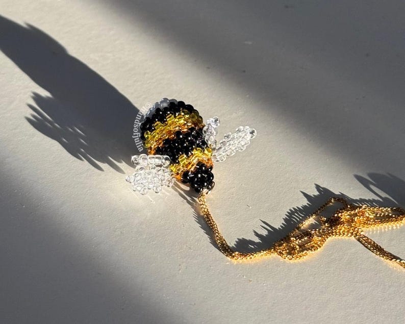 May include: A beaded bee pendant necklace. The bee is black, yellow, and clear, with a gold-colored chain. The necklace is on a white surface, with a long shadow cast by the sun.
