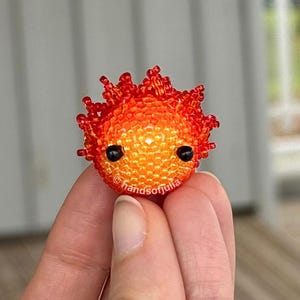 Puede incluir: Una criatura esf&eacute;rica de cuentas, con una combinaci&oacute;n de colores naranja y rojo. El trabajo de cuentas hecho a mano presenta peque&ntilde;os ojos negros y una corona puntiaguda. El texto "handsofjulia" es visible. Objeto de arte artesanal &uacute;nico.