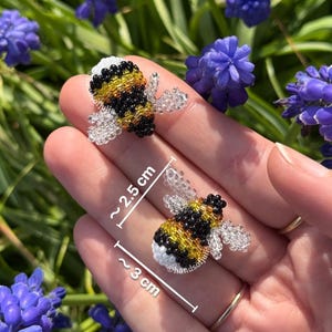 May include: Two beaded bee ornaments, approximately 1 inch and 1.2 inches in length, are displayed against a backdrop of purple flowers. The bees feature black, yellow, and white beads with clear wings.