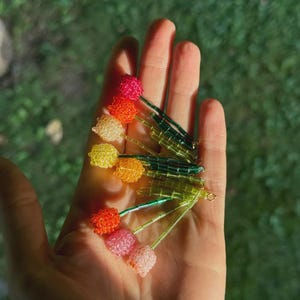 Puede incluir: Horquillas para el pelo con forma de tulipanes de cuentas en varios colores, como rosa, naranja, amarillo y rojo. Los tallos verdes se sostienen en una mano sobre un fondo verde borroso.