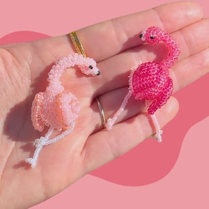 Puede incluir: Dos figuras de flamencos de cuentas en tonos rosa y blanco. Los flamencos tienen ojos negros y posan con el cuello curvado. El fondo es de color rosa liso. Las figuras est&aacute;n hechas a mano.