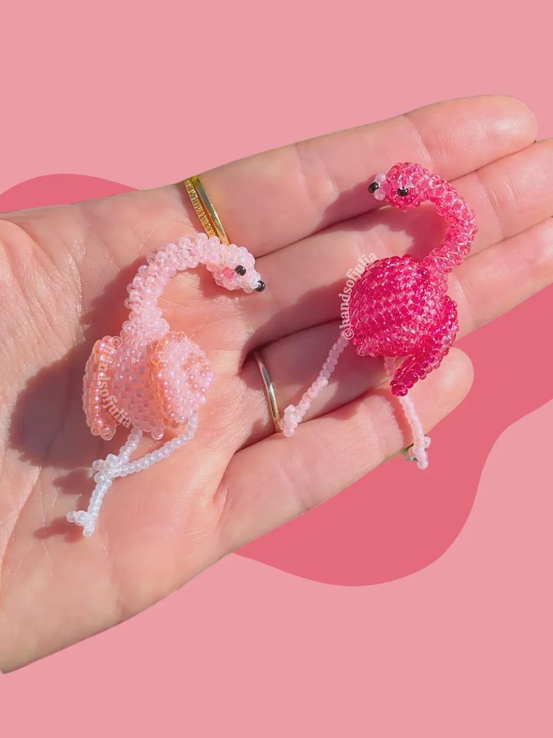 Puede incluir: Dos figuras de flamencos de cuentas en tonos rosa y blanco. Los flamencos tienen ojos negros y posan con el cuello curvado. El fondo es de color rosa liso. Las figuras est&aacute;n hechas a mano.