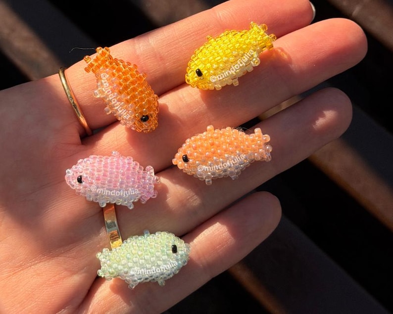 Pu&ograve; includere: Cinque anelli a forma di pesce con perline nei toni dell'arancione, giallo, rosa e verde. Ogni anello presenta un piccolo design di pesce dettagliato con occhi neri. Gli anelli sono esposti su una mano, mostrando i gioielli colorati e fatti a mano.