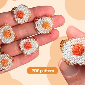 Puede incluir: Una colección de seis motivos de huevos fritos hechos con cuentas, cada uno elaborado con cuentas blancas para la clara y cuentas naranjas para la yema. Los motivos están dispuestos sobre una mano y una etiqueta naranja muestra el texto PDF pattern.