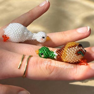 Puede incluir: Tres figuras de animales con cuentas: un ganso blanco, un pato verde y un pato marr&oacute;n. El ganso tiene patas y pico naranjas, el pato tiene cabeza verde y el pato marr&oacute;n tiene patas naranjas. Tambi&eacute;n se ve un anillo de oro.