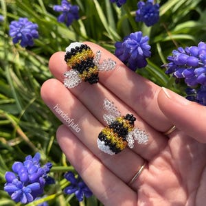 Puede incluir: Dos adornos de abeja de cuentas con cuerpos amarillos, negros y blancos y alas transparentes. Las abejas se sostienen en una mano, con un fondo de flores moradas y follaje verde. El texto "handsofjulia" es visible.
