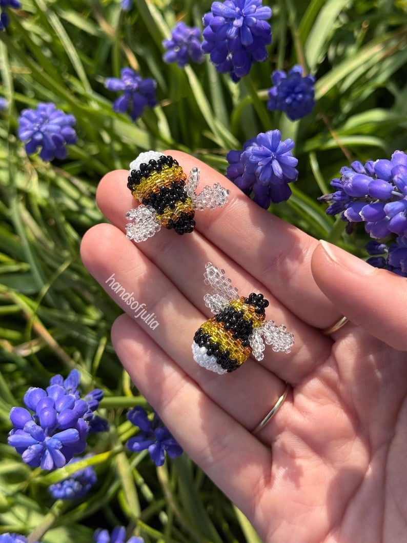 Puede incluir: Dos adornos de abeja de cuentas con cuerpos amarillos, negros y blancos y alas transparentes. Las abejas se sostienen en una mano, con un fondo de flores moradas y follaje verde. El texto "handsofjulia" es visible.
