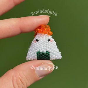 Puede incluir: Un charm de onigiri hecho a mano, con cuentas. El arroz blanco est&aacute; acentuado con cuentas naranjas y una base de algas verdes. El charm tiene ojos de cuentas negras y una cara sonriente. El texto "mindofjulia" es visible.
