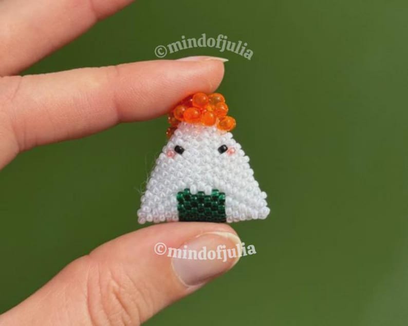 Puede incluir: Un charm de onigiri hecho a mano, con cuentas. El arroz blanco est&aacute; acentuado con cuentas naranjas y una base de algas verdes. El charm tiene ojos de cuentas negras y una cara sonriente. El texto "mindofjulia" es visible.