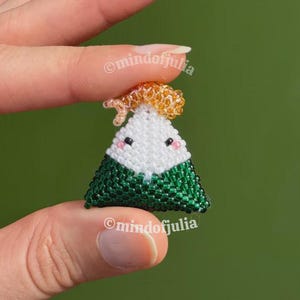 Puede incluir: Un charm de onigiri hecho a mano con cuentas. El charm es triangular, con una cara blanca, algas verdes y un peque&ntilde;o adorno naranja. El charm tiene ojos negros y mejillas rosas. El texto "@mindofjulia" es visible.