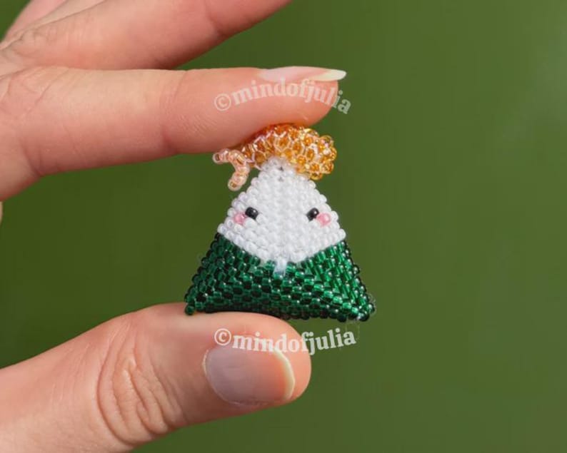 Puede incluir: Un charm de onigiri hecho a mano con cuentas. El charm es triangular, con una cara blanca, algas verdes y un peque&ntilde;o adorno naranja. El charm tiene ojos negros y mejillas rosas. El texto "@mindofjulia" es visible.