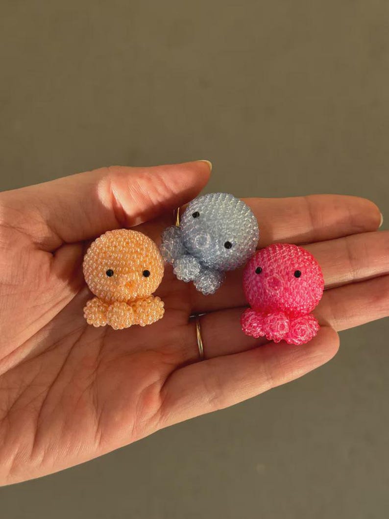 Puede incluir: Tres figuras de pulpos con cuentas en naranja, azul y rosa. Cada pulpo tiene ojos negros y un cuerpo redondeado y texturizado con tent&aacute;culos rizados. Las figuras son peque&ntilde;as y parecen hechas a mano.