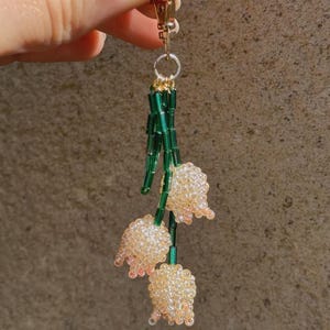 Puede incluir: Un charm de cuentas con tres flores de lirio de los valles. Las flores est&aacute;n hechas de peque&ntilde;as cuentas claras con acentos rosados, unidas a tallos de cuentas verdes. El charm tiene un cierre dorado.