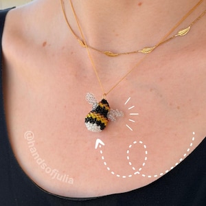 May include: A handmade bee pendant necklace. The bee pendant is black, yellow, and white with delicate wings. The necklace has a gold chain with a second chain featuring leaf-shaped charms. The necklace is worn on a person.