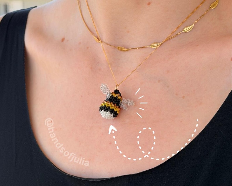 May include: A handmade bee pendant necklace. The bee pendant is black, yellow, and white with delicate wings. The necklace has a gold chain with a second chain featuring leaf-shaped charms. The necklace is worn on a person.