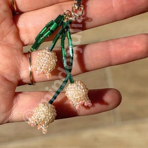 May include: A beaded keychain featuring three flower-shaped charms. The charms are made of small, light-colored beads with pink accents, attached to green beaded stems. The keychain has a gold-colored clasp.