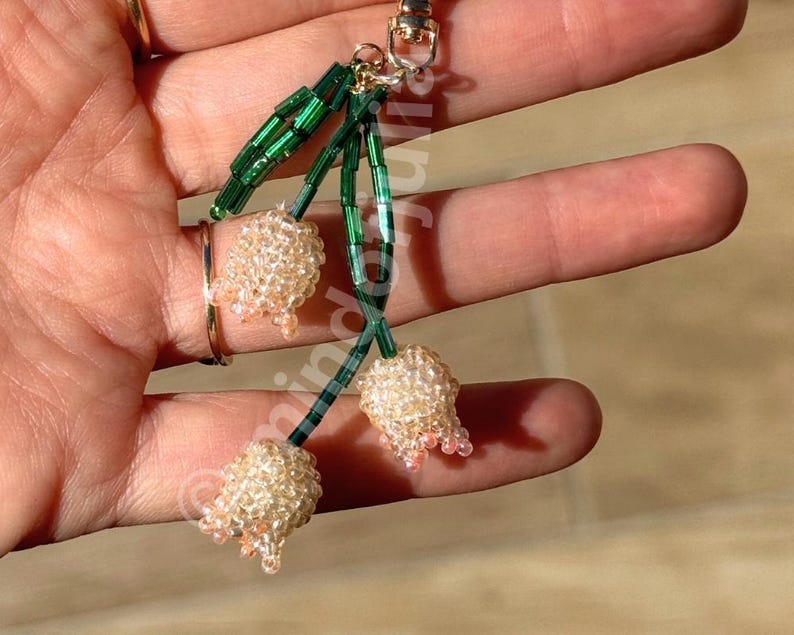 May include: A beaded keychain featuring three flower-shaped charms. The charms are made of small, light-colored beads with pink accents, attached to green beaded stems. The keychain has a gold-colored clasp.