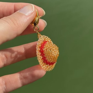 Puede incluir: Un pendiente dorado con forma de sombrero de paja peque&ntilde;o con una banda roja. El sombrero est&aacute; hecho de material tejido y tiene un cierre dorado. El fondo es verde.