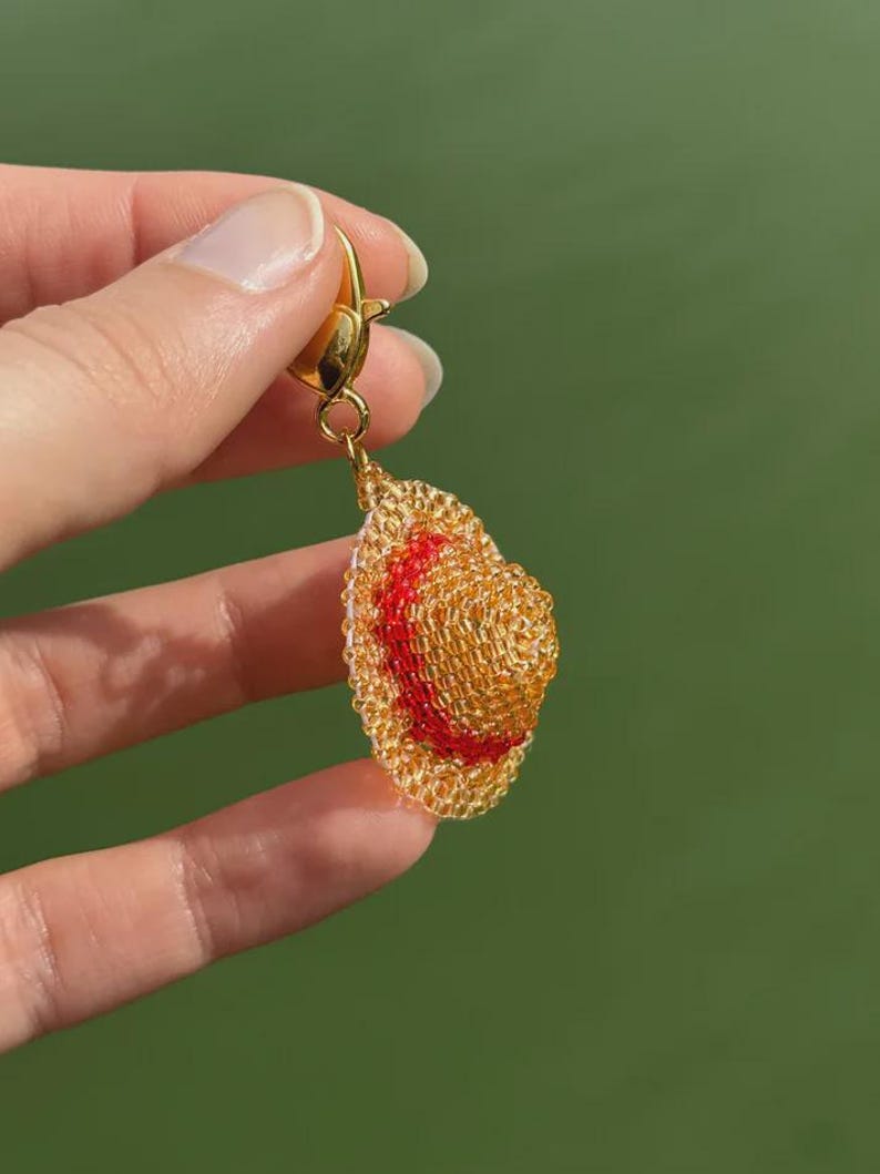 Puede incluir: Un pendiente dorado con forma de sombrero de paja peque&ntilde;o con una banda roja. El sombrero est&aacute; hecho de material tejido y tiene un cierre dorado. El fondo es verde.