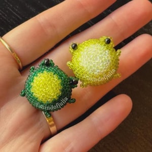 Puede incluir: Dos anillos de rana de cuentas. Uno es verde y amarillo, el otro amarillo y verde. Los anillos est&aacute;n en una banda dorada. El texto "@mindofjulia" est&aacute; en los anillos.