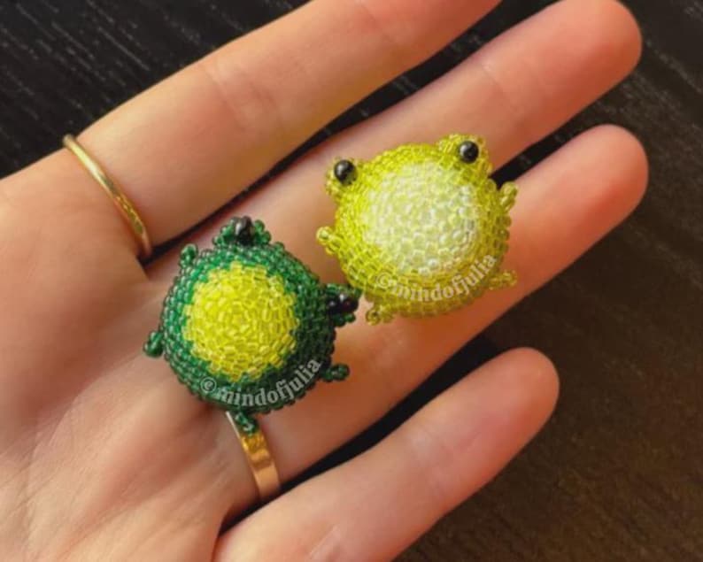 Puede incluir: Dos anillos de rana de cuentas. Uno es verde y amarillo, el otro amarillo y verde. Los anillos est&aacute;n en una banda dorada. El texto "@mindofjulia" est&aacute; en los anillos.