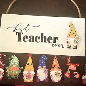 May include: A white wooden sign with the words "best Teacher ever" in black script and block letters. The sign is decorated with several gnome cutouts, each with a unique design and color scheme. A rope is attached for hanging.