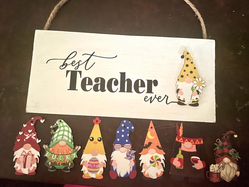 May include: A white wooden sign with the words "best Teacher ever" in black script and block letters. The sign is decorated with several gnome cutouts, each with a unique design and color scheme. A rope is attached for hanging.