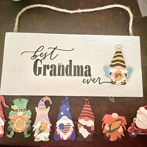 May include: A white wooden sign with the words "Best Grandma ever" in black script and block letters. The sign is decorated with several gnome cutouts, including a bee-themed gnome and others with various designs and themes.