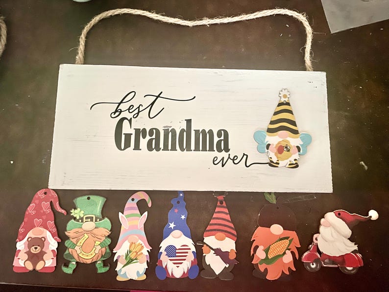 May include: A white wooden sign with the words "Best Grandma ever" in black script and block letters. The sign is decorated with several gnome cutouts, including a bee-themed gnome and others with various designs and themes.