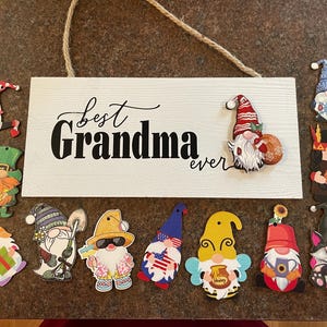 May include: A wooden sign with the words "Best Grandma ever" in black script, surrounded by colorful gnome cutouts. The gnomes have various designs, including patriotic, holiday, and floral themes.
