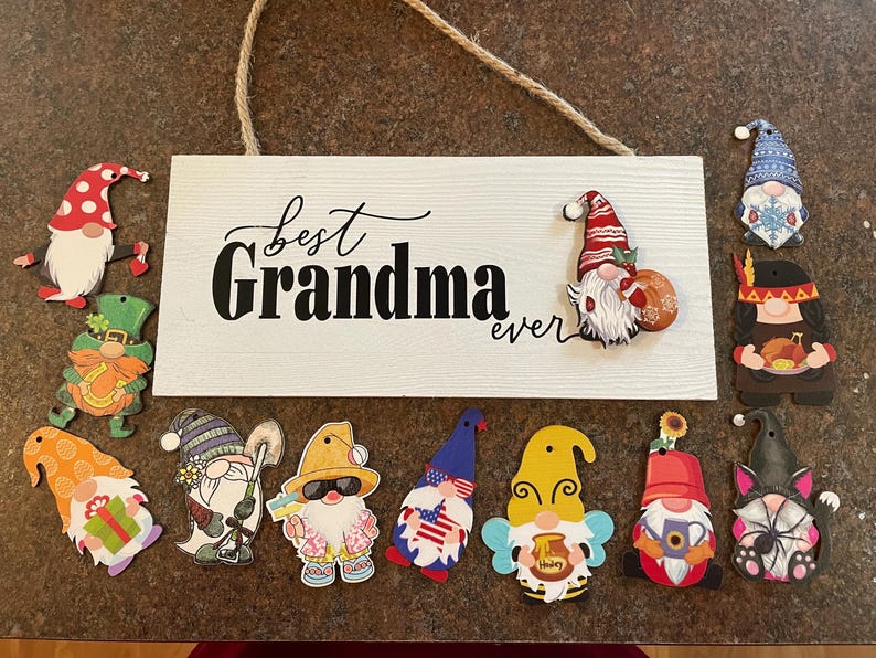 May include: A wooden sign with the words "Best Grandma ever" in black script, surrounded by colorful gnome cutouts. The gnomes have various designs, including patriotic, holiday, and floral themes.
