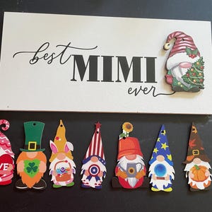 May include: A white wooden sign with the words "best MIMI ever" in black script and block letters.  A Christmas gnome is attached to the sign. Below are seven colorful gnome cutouts, each with a unique design, including hearts, shamrocks, and stars.