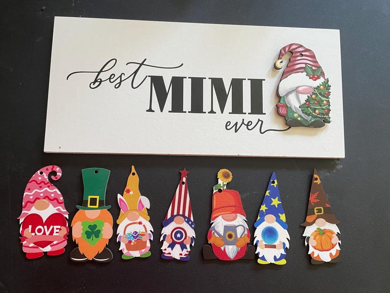 May include: A white wooden sign with the words "best MIMI ever" in black script and block letters.  A Christmas gnome is attached to the sign. Below are seven colorful gnome cutouts, each with a unique design, including hearts, shamrocks, and stars.